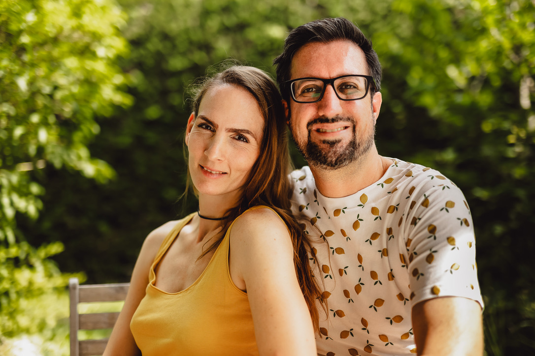 Miriam und Manuel sitzen eng nebeneinander im Grünen und lächeln gemeinsam in die Kamera