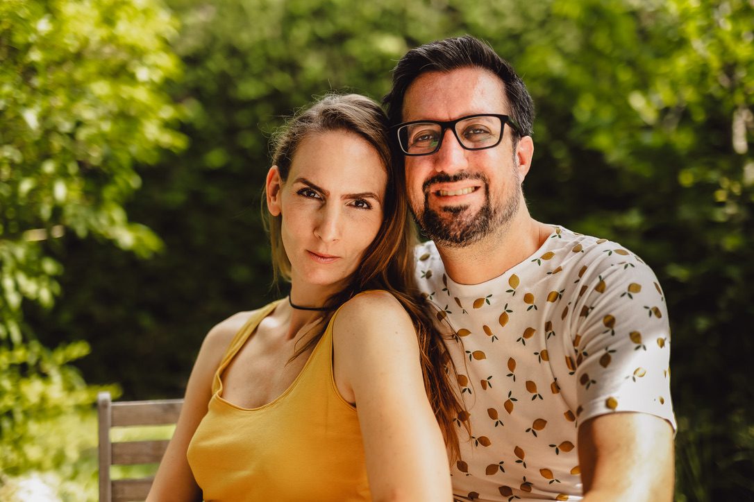 Miriam und Manuel sitzen Schulter an Schulter im Garten – Miriam mit ernstem Blick, Manuel lächelnd