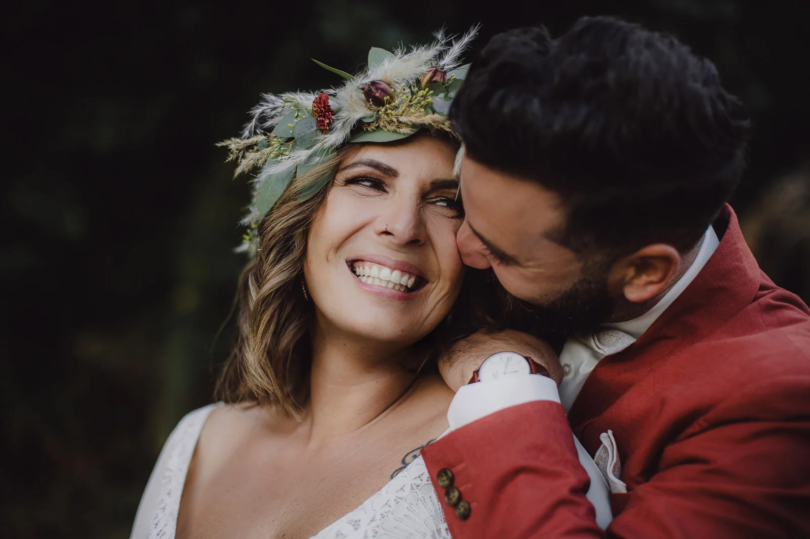 Braut mit Blumenkranz lacht, während der Bräutigam im roten Anzug sie auf die Wange küsst