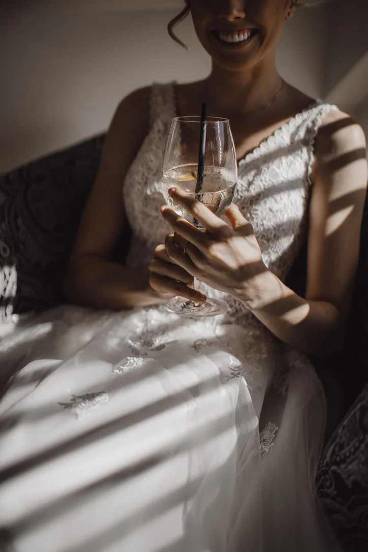 Braut sitzt im Spitzenkleid mit einem Glas in der Hand im warmen Streifenlicht am Fenster