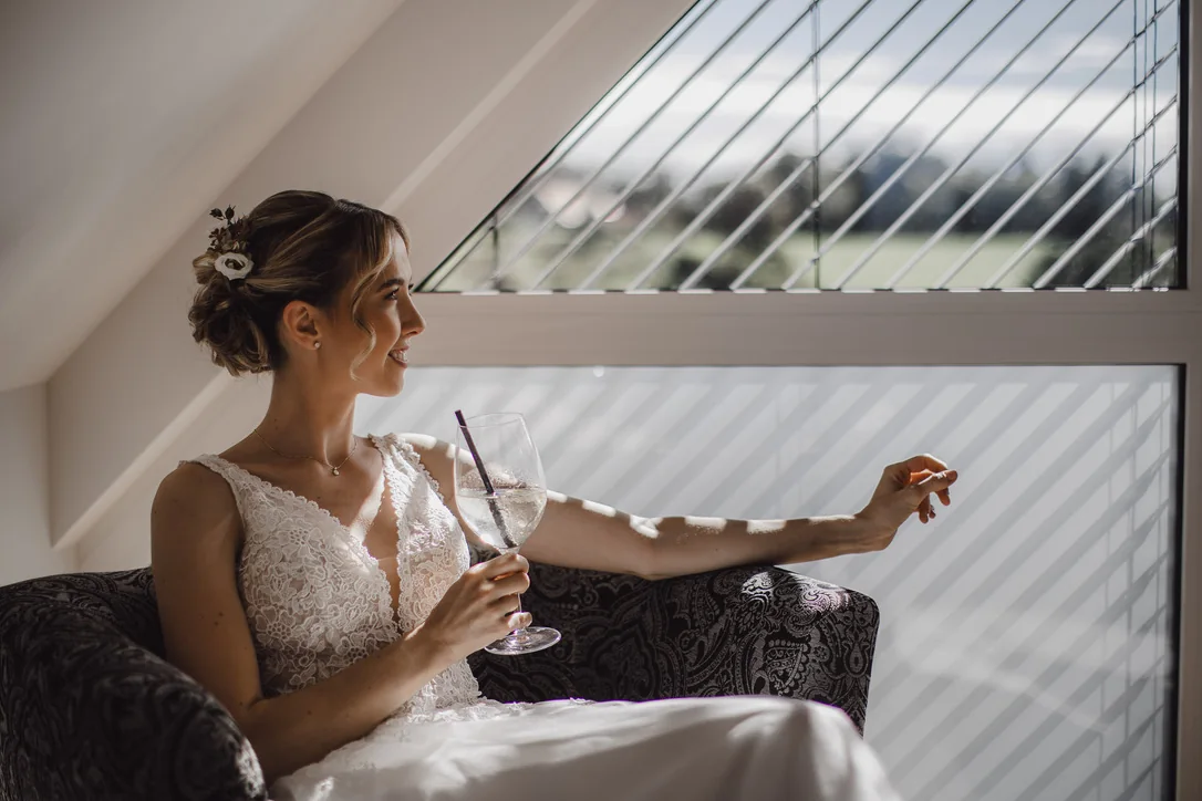 Braut sitzt entspannt mit einem Glas am großen Dachfenster und blickt lächelnd zur Seite