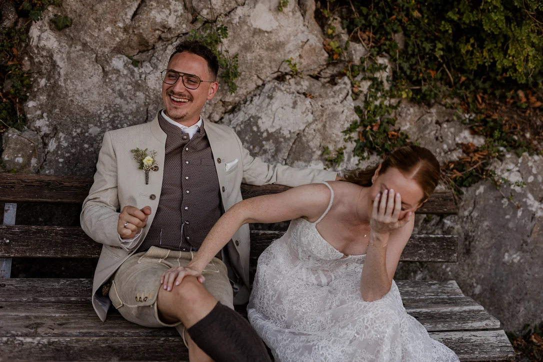 Braut und Bräutigam lachen gemeinsam auf einer Steinbank bei ihrer Hochzeit am Traunsee