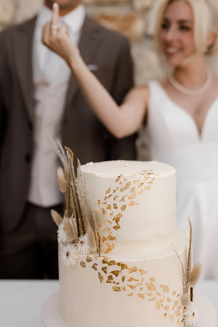 Weiße zweistöckige Hochzeitstorte mit goldener Blätterverzierung, das Brautpaar unscharf dahinter