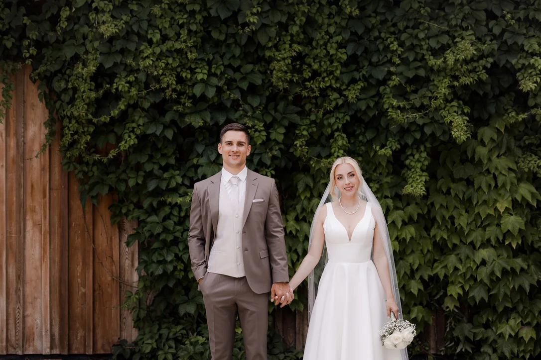 Brautpaar hält sich vor einer dicht mit Efeu bewachsenen Wand am südsteirischen Weingut an den Händen