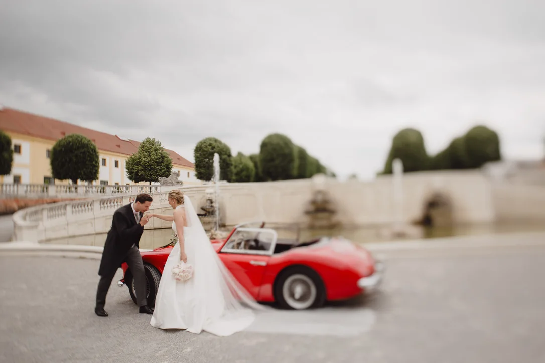 Brautpaar geht vor der barocken Schlossanlage auf einen roten klassischen Roadster zu