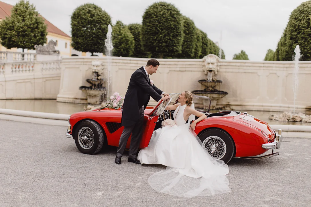 Bräutigam hilft der Braut beim Einsteigen in einen roten klassischen Roadster vor dem Schloss