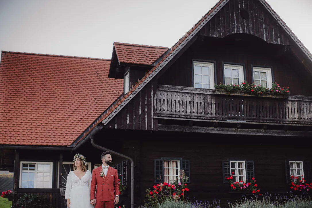 Brautpaar steht vor einem dunklen Holzhaus mit rotem Dach bei der Familienhochzeit am Weingut