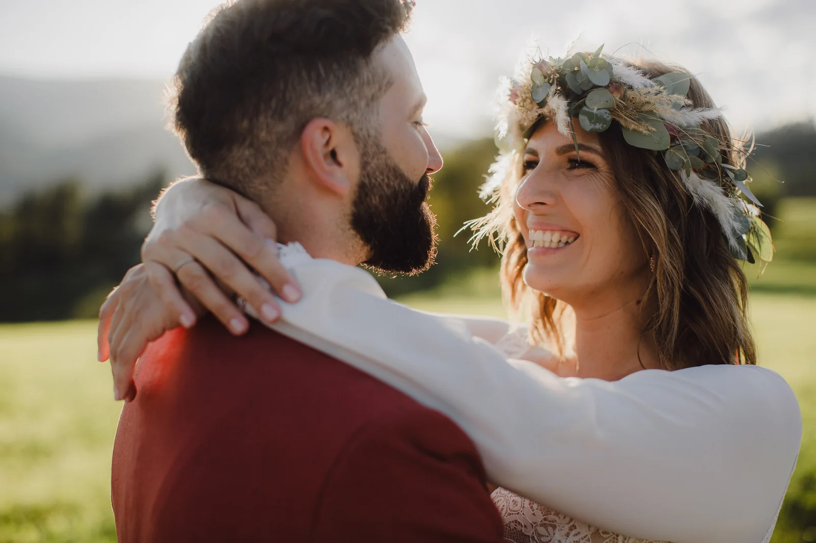 Braut mit Blumenkranz umarmt ihren Bräutigam im roten Anzug im warmen Gegenlicht der Abendsonne