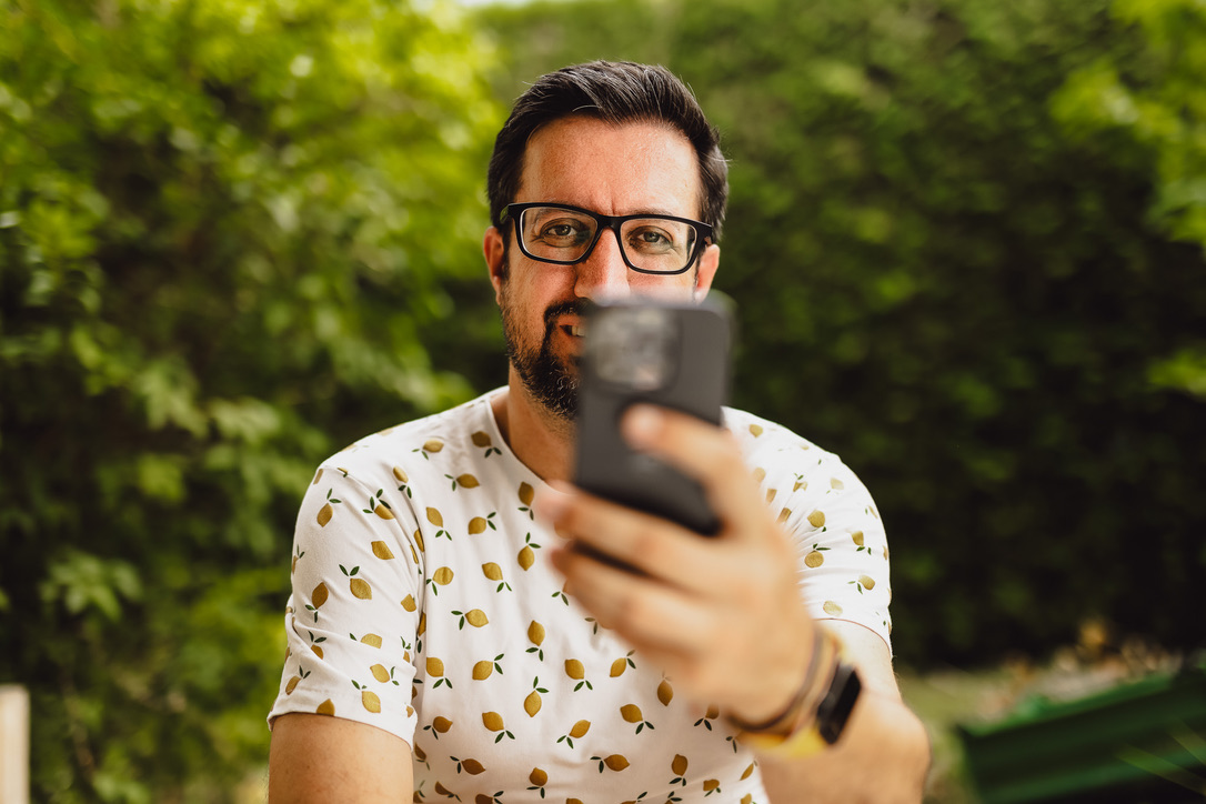 Manuel hält beim Sitzen im Garten ein Smartphone in Richtung Kamera