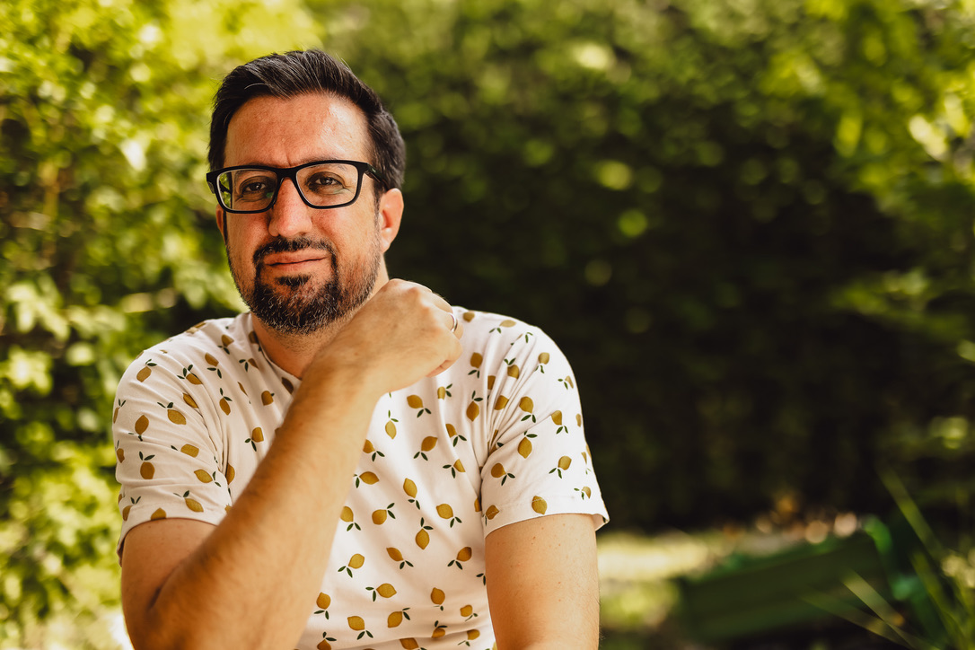 Manuel sitzt entspannt im Garten, stützt die Hand an den Hals und blickt in die Kamera