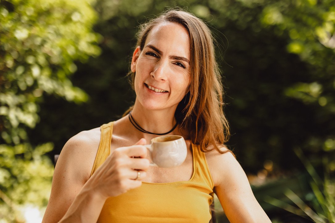 Miriam lächelt mit einer kleinen Kaffeetasse in der Hand direkt in die Kamera